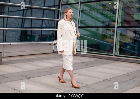 Bella donna bionda incinta con un abito da lavoro bianco con un blazer e un vestito, che cammina all'aperto con un laptop vicino a un edificio aziendale Foto Stock