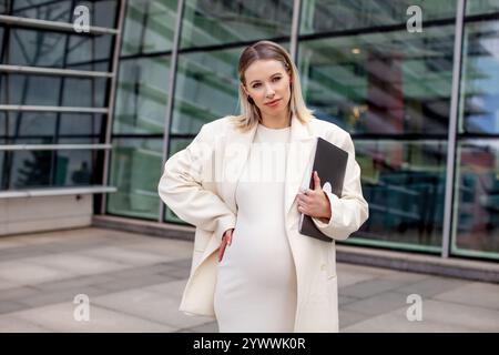 Una bella donna bionda incinta con un abito da lavoro bianco e un blazer si trova all'aperto con un notebook vicino a un edificio aziendale, che mostra una professionista Foto Stock