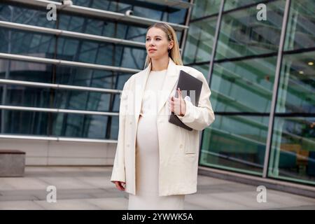 Una bella donna d'affari bionda incinta con un abito da lavoro bianco e un blazer si trova all'aperto con un notebook vicino a un edificio aziendale, che mostra un professionista Foto Stock