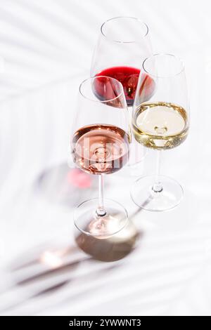 Tre bicchieri da vino pieni di vino rosso, bianco e rosa, in piedi graziosamente su uno sfondo luminoso e minimalista. Una vivace esposizione di varietà di vini Foto Stock