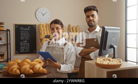 Uomo e donna in piedi insieme in una panetteria, indossando grembiuli e sorridenti, con pane fresco e pasticcini sullo sfondo e un tablet in mano Foto Stock