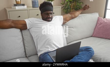 Un bel giovane afroamericano con la barba si allunga su un divano nel suo salotto mentre utilizza un computer portatile, catturando un momento di relax per adulti Foto Stock