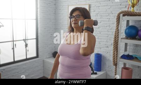 Donna ispanica di mezza età che solleva un manubrio in palestra, indossa abiti da ginnastica, pratica allenamento di forza al chiuso con attrezzature sportive Foto Stock