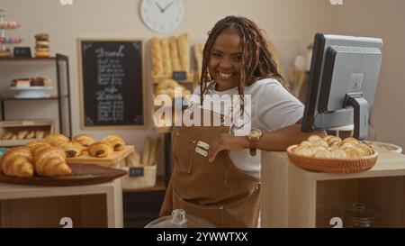 Donna sorridente in una panetteria con pasticcini e pane dietro il bancone in un negozio interno con interni in legno, orologio sulla parete e cassa Foto Stock