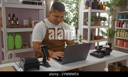 Giovane che lavora in un negozio di panetteria, utilizza un computer portatile, circondato da prodotti e oggetti decorativi sugli scaffali, con piante che aggiungono un'atmosfera fresca al Foto Stock