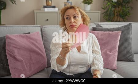 Una bella donna bionda è seduta su un divano in un soggiorno mentre si lancia, raffigurando una donna caucasica di mezza età in un ambiente domestico. Foto Stock