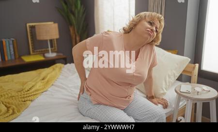 Un'anziana donna caucasica siede sul suo letto in una camera da letto, mostrando disagio nella schiena, in casa. Foto Stock