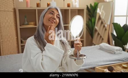 Donna matura che si rilassa in una serena camera spa, si ammira in uno specchio con un sorriso premuroso, mettendo in risalto l'autocura e la bellezza in un benessere Foto Stock