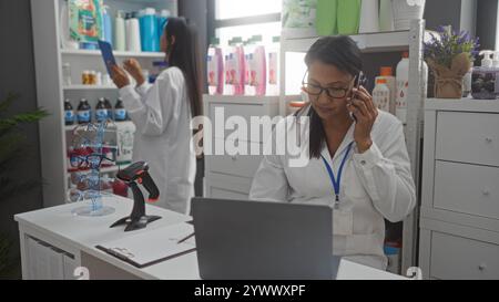 Due farmacie femminili in una farmacia che lavorano insieme, una al telefono, mentre l'altra utilizza un tablet, circondato da scaffali con vari sanitari p Foto Stock