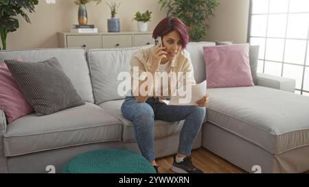 Una giovane donna con i capelli corti è seduta nel suo salotto, parla al telefono mentre tiene i documenti, il che indica un ambiente domestico sereno in un W Foto Stock