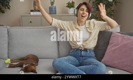 La giovane donna con le cuffie balla gioiosamente su un divano in un accogliente soggiorno con il suo cane che dorme accanto a lei. Foto Stock