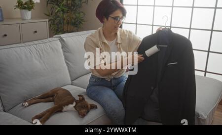 Una giovane donna che usa un rullo di lanugine su una giacca nera mentre siede su un divano con il suo cane in un elegante soggiorno. Foto Stock