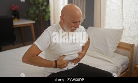 Uomo che soffre di dolore al petto seduto a letto in camera da letto con le mani che stringono lo stomaco mostrando disagio Foto Stock