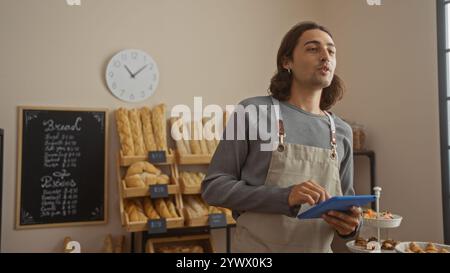 Giovane ispanico con lunghi capelli e grembiule che tiene la tavoletta in una panetteria circondata da scaffali di pane e dolci. Foto Stock