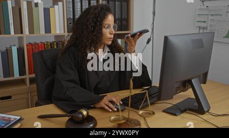 Una giovane donna giudica con i capelli ricci che parla al telefono seduto alla scrivania in ufficio con computer e oggetti legali Foto Stock