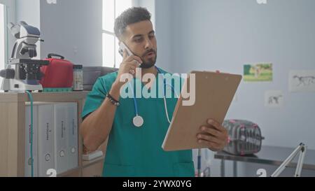Un bel giovane ispanico in una clinica veterinaria esamina un appunti mentre parla al telefono, indossando scrub verdi e uno stetoscopio intorno al suo Foto Stock