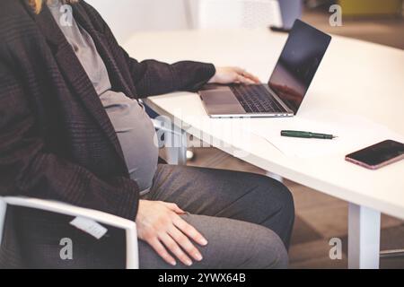 Primo piano di una donna incinta che lavora su un notebook con un desktop bianco. Donna d'affari incinta corta che scrive sul laptop mentre è seduta al suo posto di lavoro in offi Foto Stock