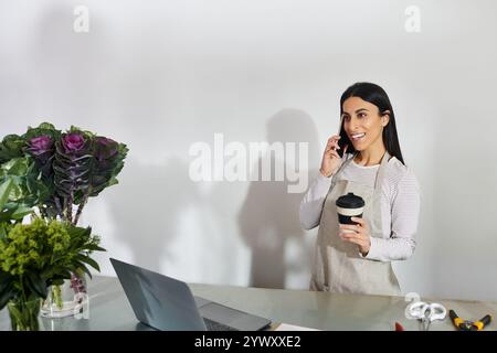 Una giovane donna gestisce il suo negozio di fiori, parla al telefono e si gusta un caffè. Foto Stock