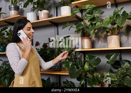 Una giovane fiorista organizza felicemente le piante mentre parla al telefono nel suo negozio. Foto Stock
