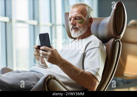 Immagine media di un uomo d'affari maturo che si prende cura della pelle del viso con le macchie per gli occhi, mentre messaggi di testo sul telefono cellulare su un bicchiere di vino rosso in una comoda poltrona a casa Foto Stock