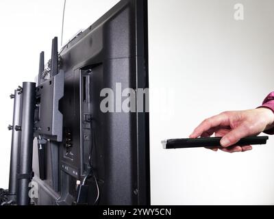 Mano di un uomo che tiene e preme un telecomando guardando il televisore, con la vista principale del soggetto sul retro del televisore su un piedistallo. Foto Stock