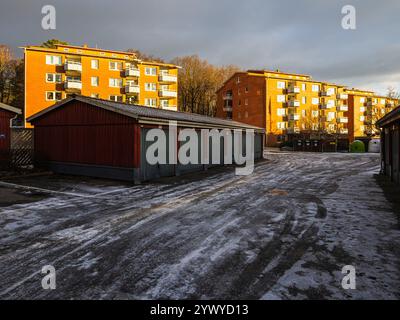 I garage fiancheggiano un sentiero innevato in un quartiere residenziale. Gli appartamenti si ergono alti sullo sfondo mentre il sole attraversa nuvole scure, creat Foto Stock