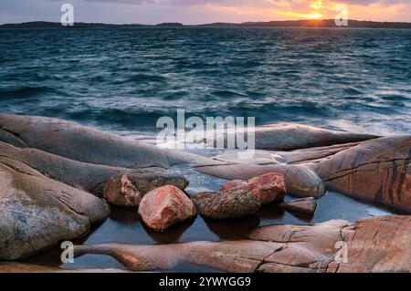 I colori caldi riempiono il cielo mentre il sole tramonta su un oceano tranquillo, gettando un bagliore dorato sulle aspre rocce lungo la riva. Le onde si infrangono dolcemente verso lo sto Foto Stock