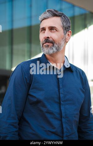 Verticale. Uomo maturo e sicuro di sé con barba grigia che guarda attentamente in lontananza, indossa una camicia blu navy, si trova all'aperto vicino a un moderno edificio in vetro, riflettendo professionalità e tranquillità. Foto di alta qualità Foto Stock