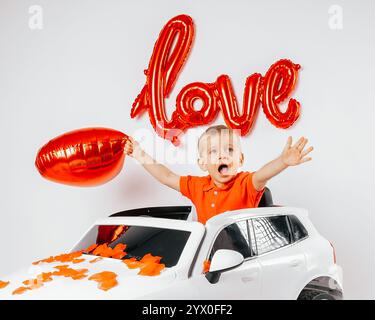 Un ragazzo biondo siede su una sedia circondato da palloncini a forma di cuore con la parola amore scritta sul muro. La scena trasuda un caldo e festoso San Valentino Foto Stock