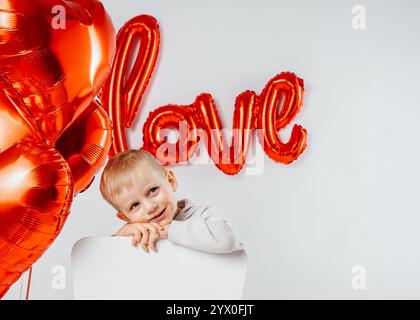 Un ragazzo biondo siede su una sedia circondato da palloncini a forma di cuore con la parola amore scritta sul muro. La scena trasuda un caldo e festoso San Valentino Foto Stock