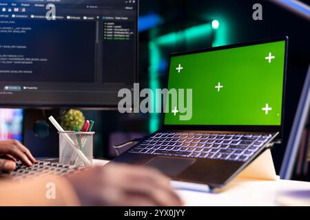 Programmatore afroamericano che lavora sul cloud computing accanto allo schermo verde, scrivendo codice per l'ottimizzazione sulla finestra del terminale. Il tecnico IT lavora per inserire codice html e sicurezza del server. Foto Stock