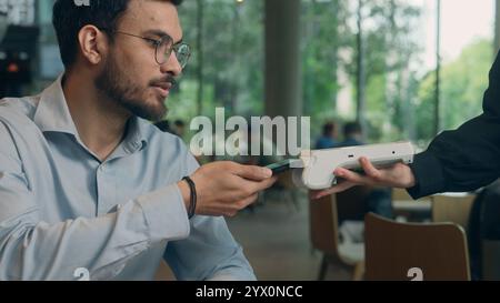 Primo piano, il visitatore paga la bolletta dell'ordine del cibo nel bar ristorante un cameriere irriconoscibile che tiene in mano la scansione wi-fi del terminale utilizzando la tecnologia NFC senza contatto dello smartphone Foto Stock