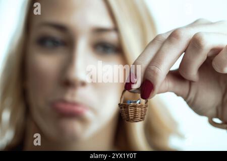 Una donna bionda con smalto rosso ha in mano un cestino in vimini contenente piccoli pacchetti regalo, il suo viso sfocato esprime delusione e dissanguamento Foto Stock
