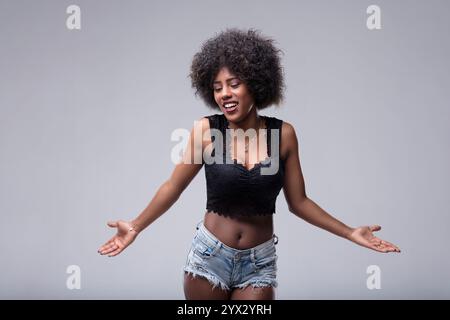 La giovane donna con un taglio di capelli afro si stringe le spalle con le braccia aperte e un'espressione confusa, guardando in basso e lateralmente contro una bara grigia Foto Stock