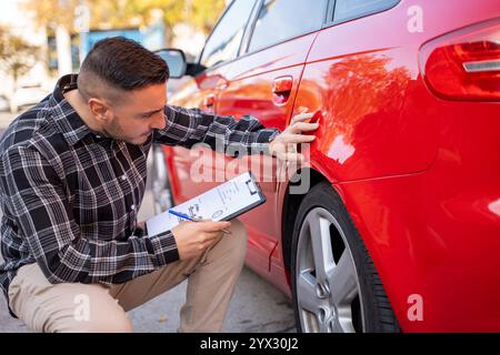 Agente assicurativo accovacciato accanto a un'auto rossa danneggiata, scrivendo appunti su un appunti per il rapporto dell'incidente Foto Stock