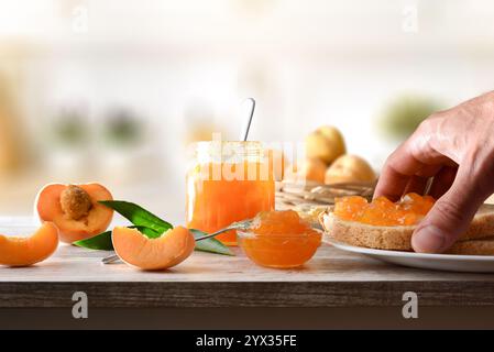 Raccogliere a mano il pane tostato con marmellata di pesca su banco di legno con contenitori con marmellata e pezzi e fette di frutta in un recipiente di frutta e in cucina sullo sfondo. Foto Stock