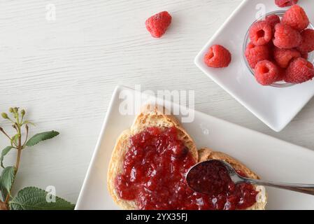 Tosta con marmellata di lamponi su un piatto bianco preparato al momento per la colazione e una ciotola piena di frutti di bosco su una panca di legno bianca. Vista dall'alto. Foto Stock