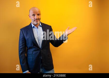 Un uomo con una tuta blu scuro punta a destra. Guarda la telecamera con un leggero sorriso. Lo sfondo è giallo fisso. Foto Stock