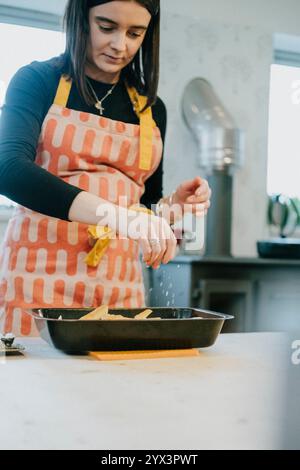 Una donna sta insaporendo il cibo in una cucina. Indossa un grembiule e una collana. La cucina è ben attrezzata con lavandino, frigorifero e vaschetta Foto Stock