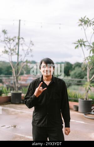 Un uomo del sud-est asiatico che fa un segno di metallo con le dita, esprimendo fiducia ed energia. Ideale per musica, stile di vita o progetti culturali Foto Stock