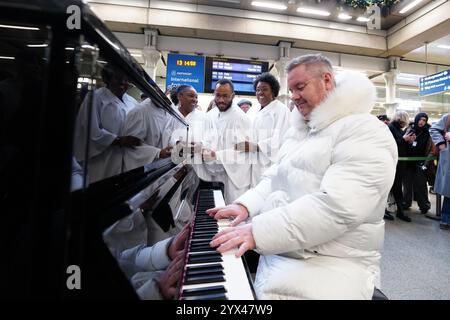 L'ex cantante degli East 17 Tony Mortimer sorprende i pendolari della stazione St Pancras International di Londra con una performance pop-up del suo numero uno natalizio 1994 "Stay Another Day", supportato dal London Community Gospel Choir. La performance arriva quando Tony ha collaborato con l'associazione benefica Nordoff e Robbins per celebrare il 30° anniversario dell'uscita della canzone. Data foto: Venerdì 13 dicembre 2024. Foto Stock