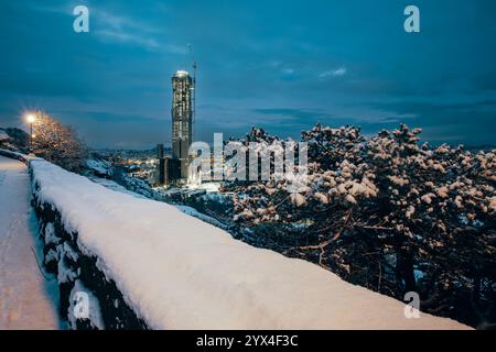 La neve ricopre il terreno e gli alberi in una città che si affaccia su un'alta torre, mentre i lampioni proiettano un caldo bagliore contro un cielo crepuscolo, creando un vento sereno Foto Stock