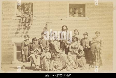 Gruppo scolastico nativo americano, circa 1870-1879. Un ritratto di gruppo di un gruppo scolastico di nativi americani. Un uomo che tiene un libro, presumibilmente l'insegnante, si trova al centro del gruppo. I bambini guardano fuori dalle finestre nell'edificio in mattoni dietro il Foto Stock