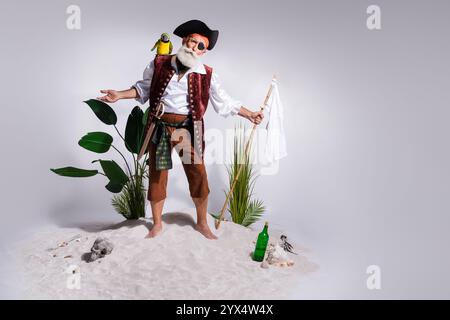 L'anziano capitano pirata con pappagallo si erge sulla spiaggia sabbiosa, reggendo una bandiera bianca, vestito con un autentico costume da bucaniere. Foto Stock