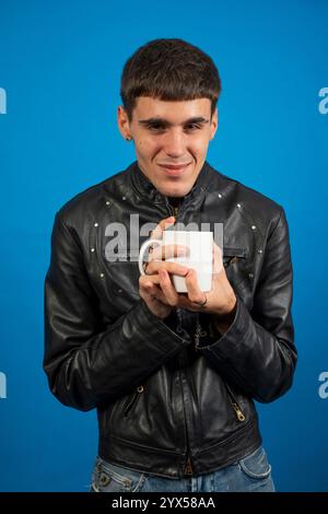 Un uomo elegante che sorseggia una bevanda calda in una tazza bianca, su un vivace sfondo blu Foto Stock