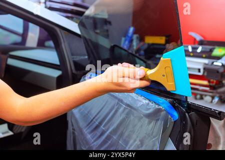 Il lavoratore a mano applica con cautela il tergivetro per montare la pellicola sul finestrino laterale del veicolo in officina. Foto Stock