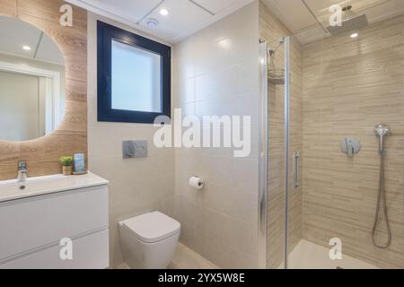 Bagno moderno con elegante cabina doccia, lavandino minimalista con specchio rotondo e luce naturale dalla finestra. Foto Stock