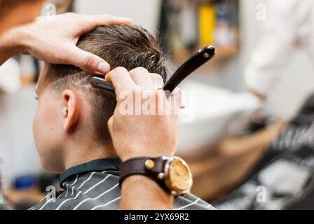 Le mani maschili tengono il rasoio dritto vicino alla testa del bambino. Taglio di capelli da uomo in barbiere. Accessori per capelli e cura dei capelli. Salone di bellezza Foto Stock