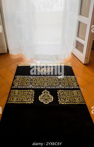 Tappeto di preghiera nero islamico con decorazioni dorate, tappeto sul pavimento di fronte ad una finestra con tende bianche, atmosfera spirituale, vibrazioni Ramadan, sfondo Foto Stock