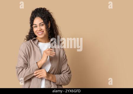 Graziosa giovane donna afroamericana che abbottonava un'elegante camicia su sfondo beige Foto Stock